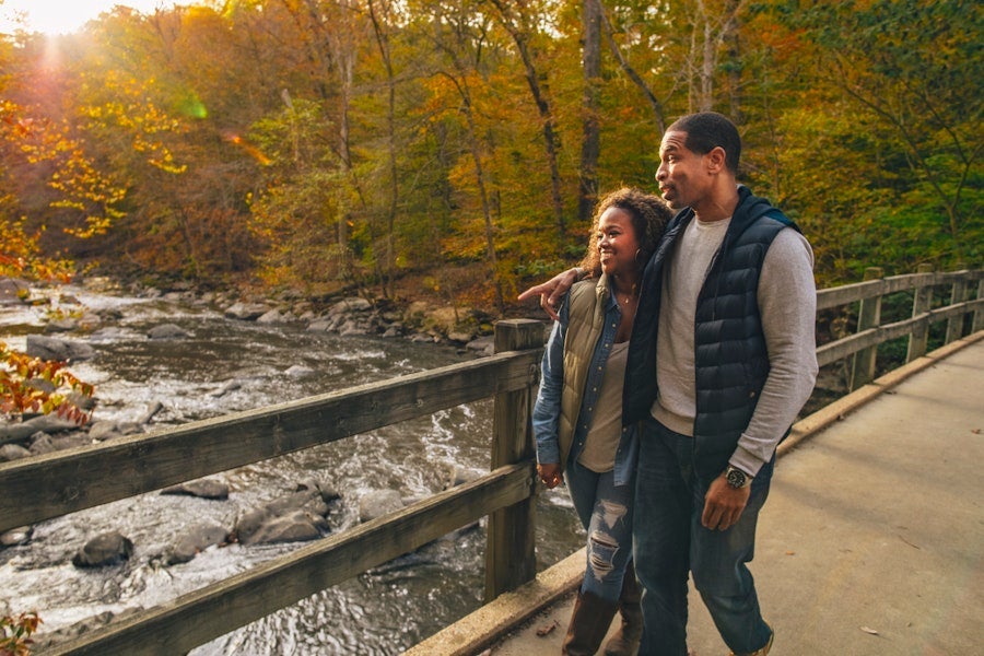 A couple walking on a bridge over a scenic river with colorful autumn foliage.