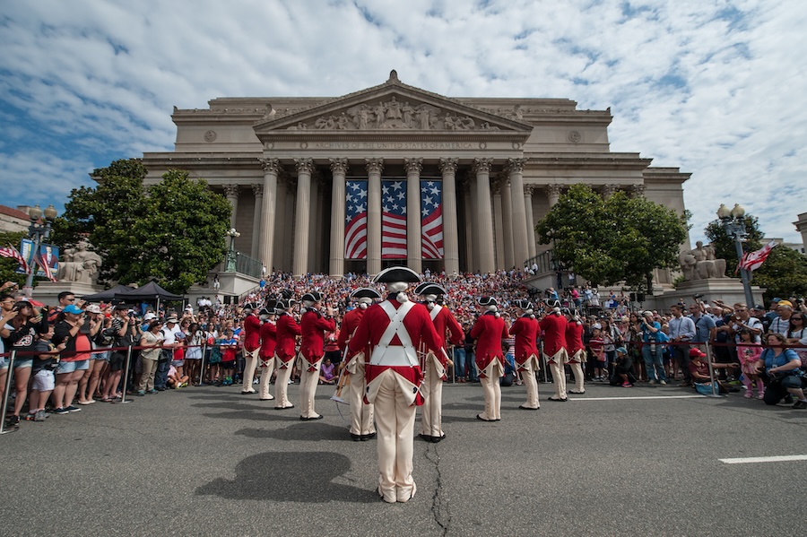 A fife and drum corps dressed in red coats performs in front of a large crowd gathered outside the National Archives, which is adorned with American flags.