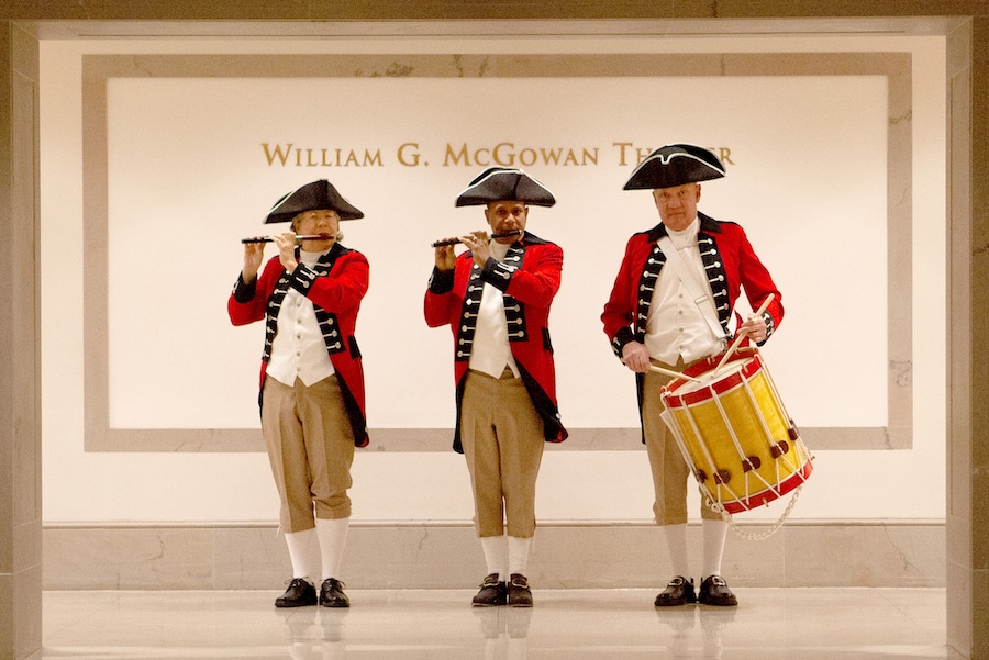 Three musicians in colonial attire play fifes and a drum inside the National Archives, standing against a backdrop with "William G. McGowan Theater" inscribed on the wall.