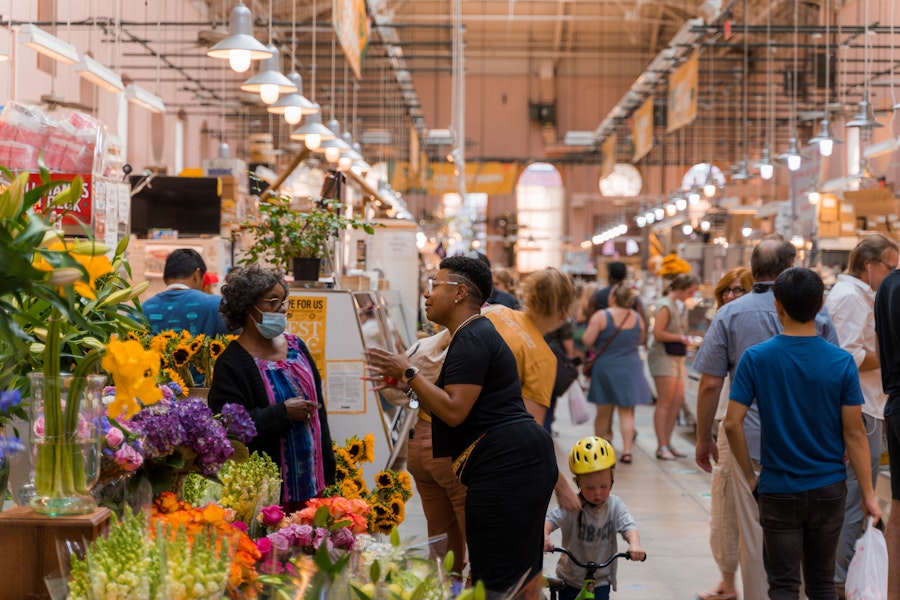 A bustling flower and produce aisle at Eastern Market, filled with shoppers, vendors and colorful blooms under bright lights.