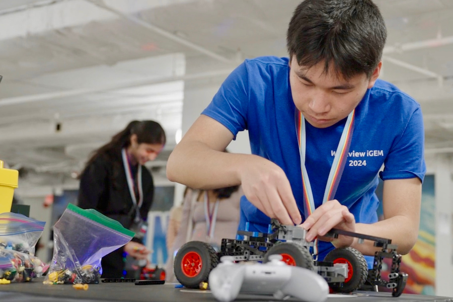 A student in a blue "Westview iGEM 2024" shirt intently assembles a wheeled robot at a STEM competition.