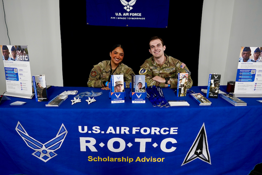 Two U.S. Air Force ROTC cadets sit smiling behind a recruitment table covered with brochures, lanyards and promotional items.