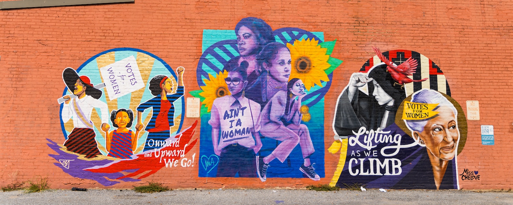 Colorful mural on a red brick wall in Anacostia celebrating women’s suffrage and empowerment. The artwork features historical and modern women of color, sunflowers, protest signs with messages like “Votes for Women” and “Ain’t I a Woman."