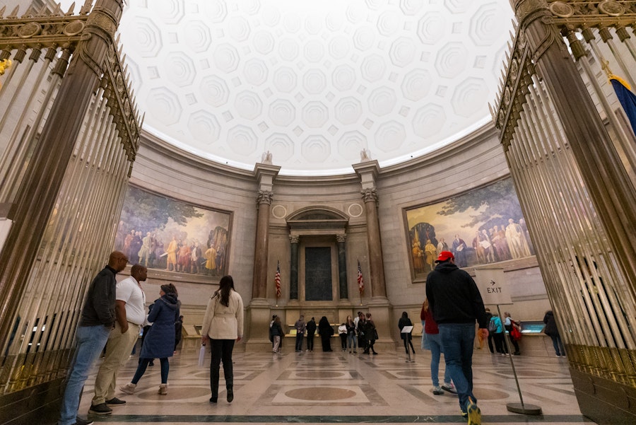 Visitors inside the National Archives Rotunda view the founding documents of the United States beneath a grand domed ceiling and large historic murals.