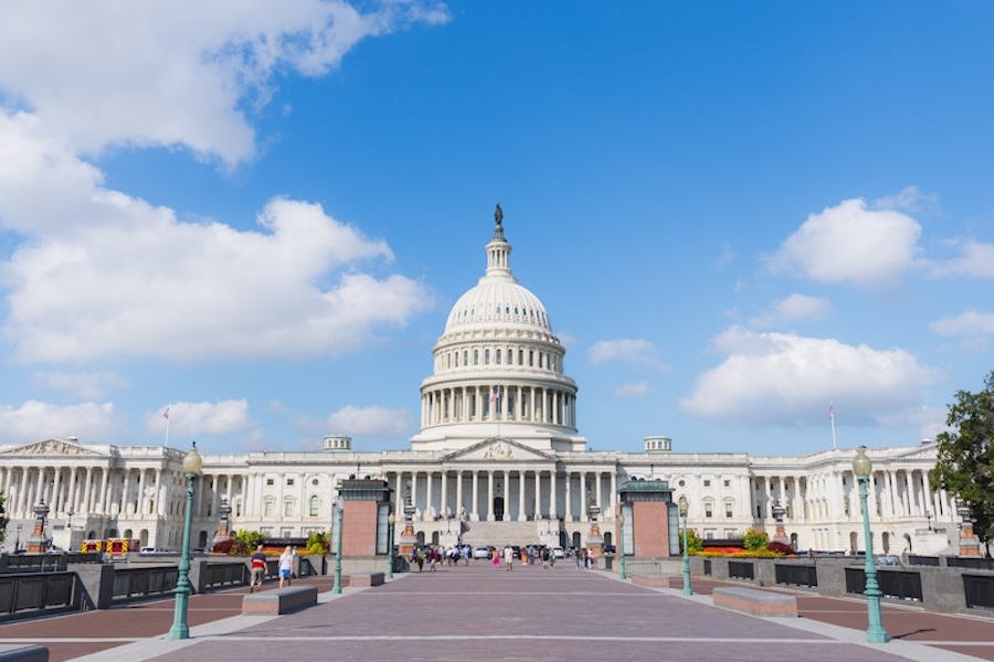 The U.S. Capitol building stands under a bright blue sky with scattered clouds, viewed from the east plaza with tourists walking nearby.