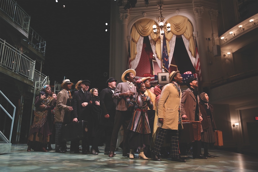 Actors perform a dramatic scene in period costumes on the historic stage of Ford’s Theatre, with the presidential box and American flags in the background.