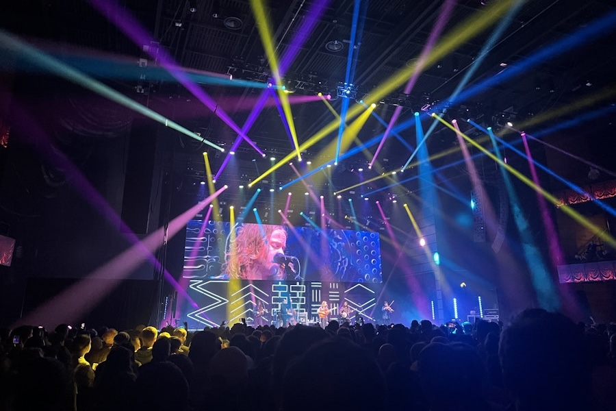 A packed crowd enjoys a live concert under a dazzling array of multicolored lights with a close-up of the performer projected on a large screen.