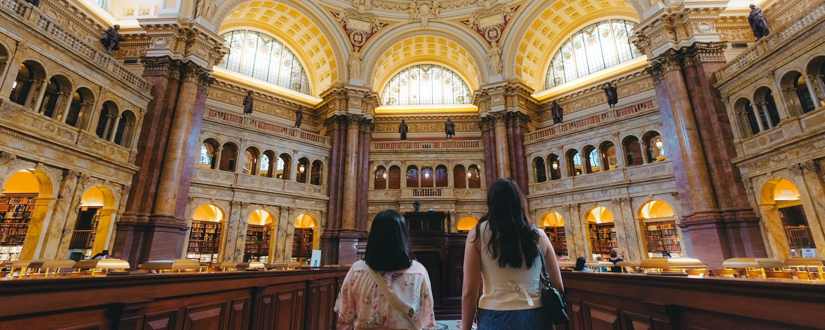 Two people gaze out into the Library of Congress' Reading Room, with grand arches and architecture. 