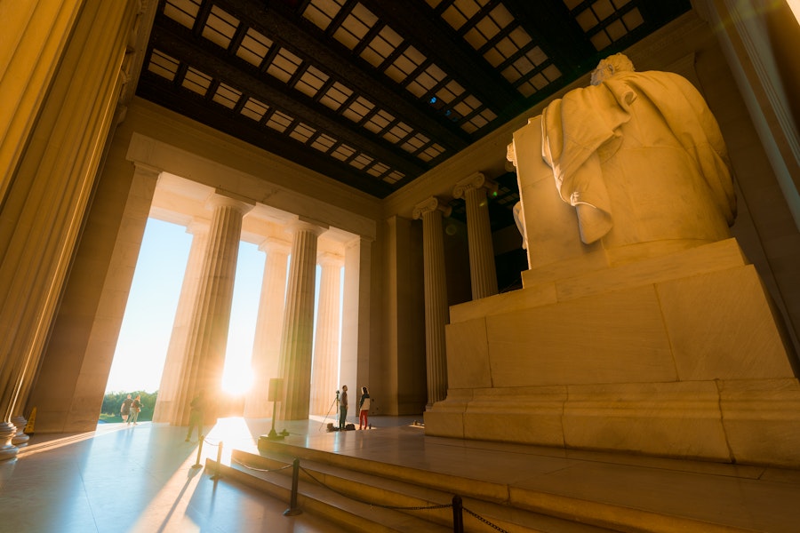 Warm sunlight pours through the columns of the Lincoln Memorial as visitors stand near the towering statue inside the historic monument.