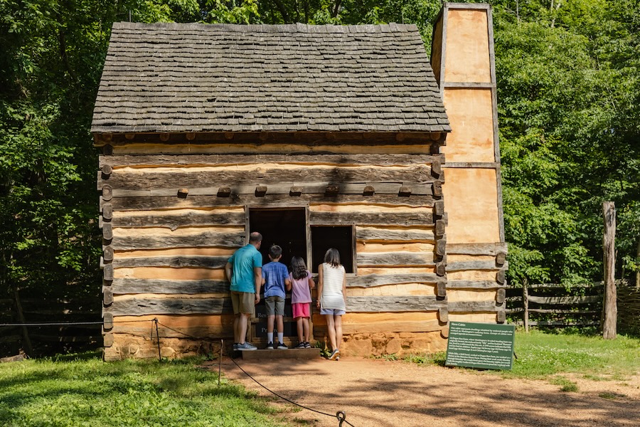 A family of four peers into a reconstructed cabin at George Washington’s Mount Vernon, surrounded by lush green trees and accompanied by an informational sign.