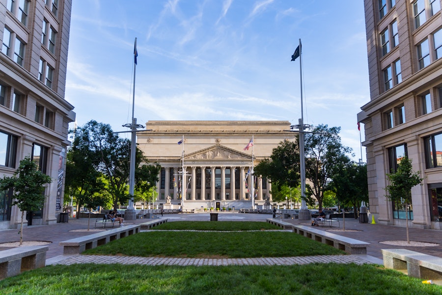 The stately facade of the National Archives building is framed between two buildings and centered behind a small plaza with flags and greenery.