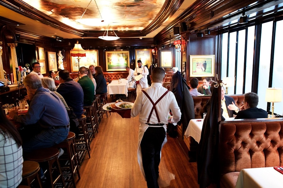 A server carries food through the dimly lit, classic interior of Old Ebbitt Grill, filled with patrons seated at leather booths and the bustling bar.