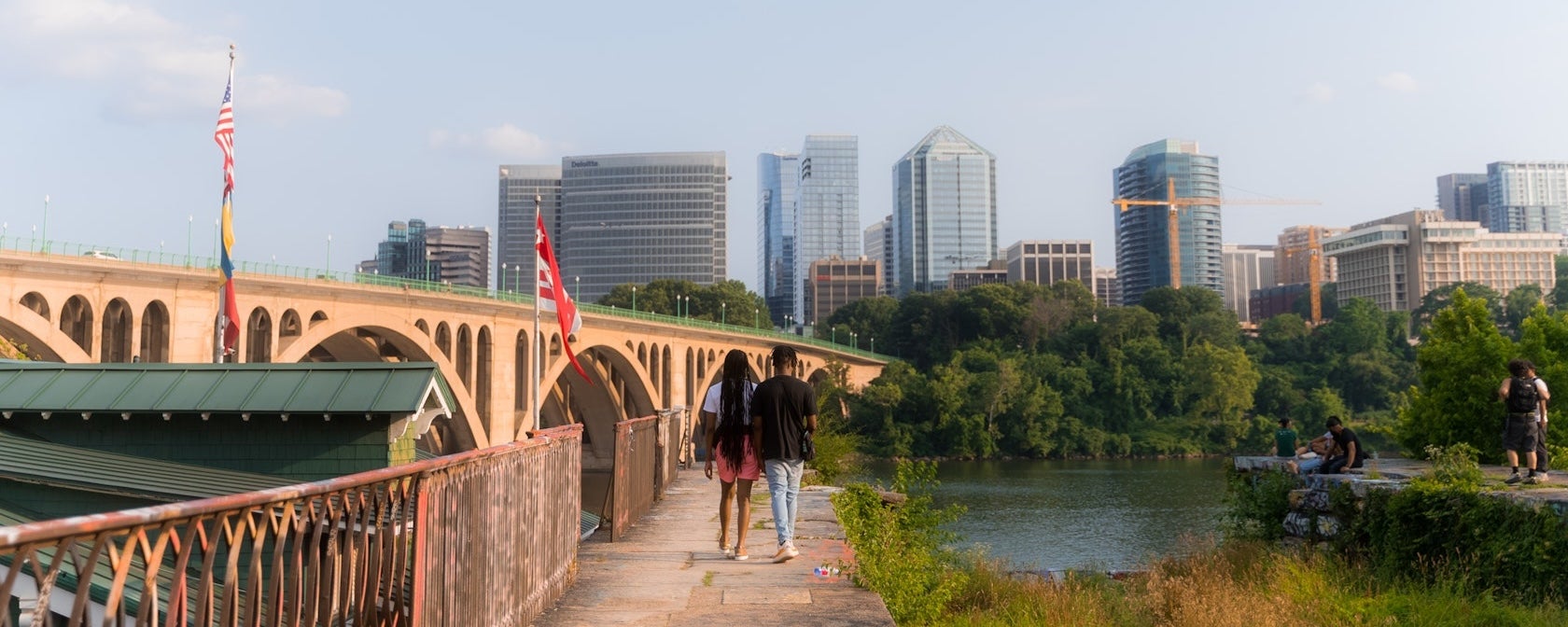 A couple walks along the Georgetown riverfront with a bridge and building of Arlington visible on the other side of the river. 