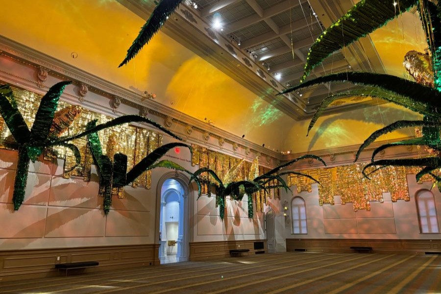 A room with golden-yellow walls and high ceilings features dramatic hanging plant-like sculptures suspended above arched doorways and murals.