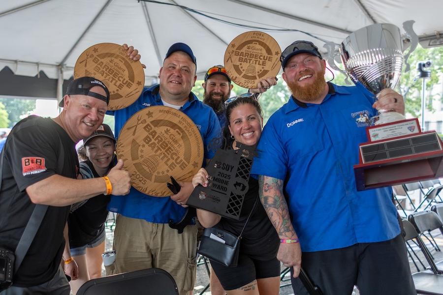 Smiling barbecue team members hold trophies and awards after a competition victory.