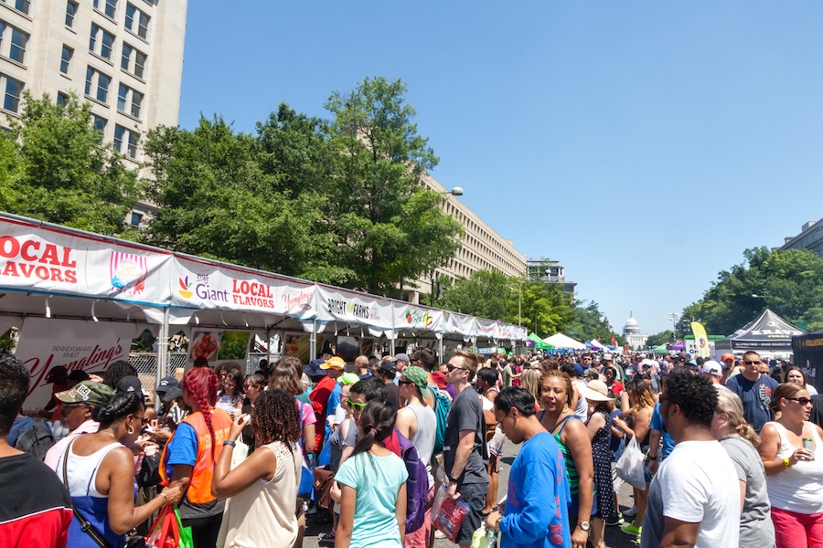 A large crowd samples food from vendor tents lined along a busy festival street.