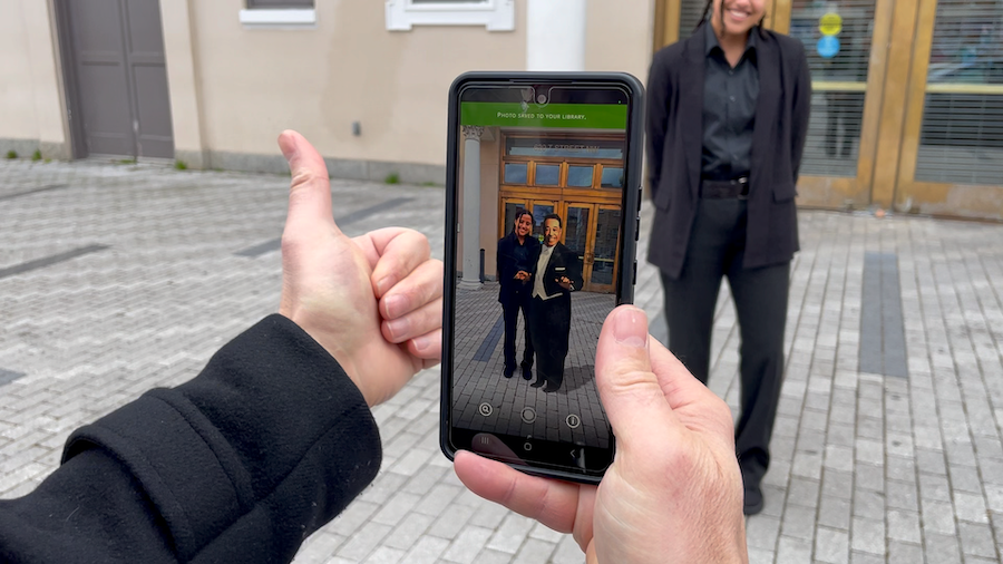 A person holds a smartphone showing an augmented reality photo of two people while another person stands smiling in the background.51-steps-augmented-reality-photo-capture-dc