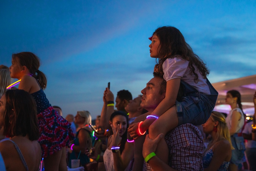 A child sits on an adult’s shoulders while watching a nighttime fireworks show with a glowing crowd.