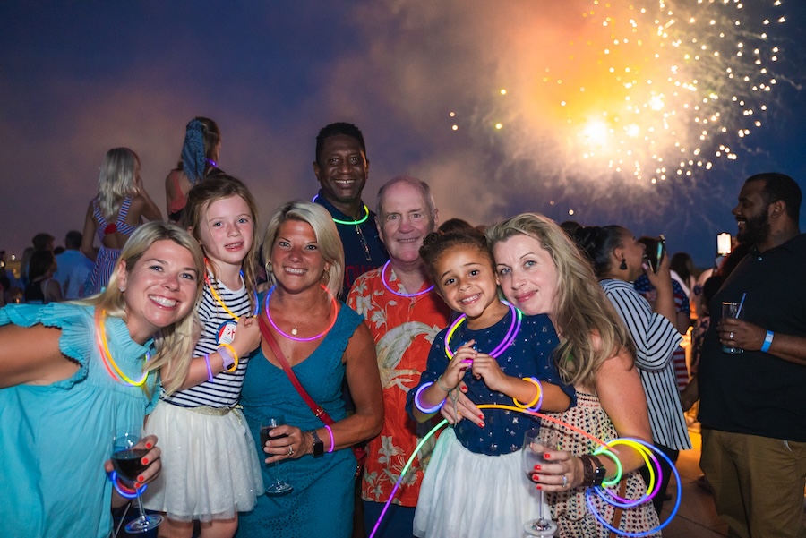 A group of adults and children smile together as fireworks light up the sky behind them.