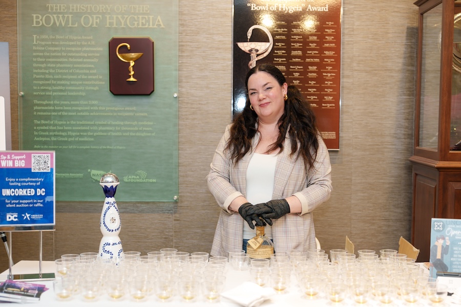 A woman stands behind a tasting table with small cups and bottles at an event.