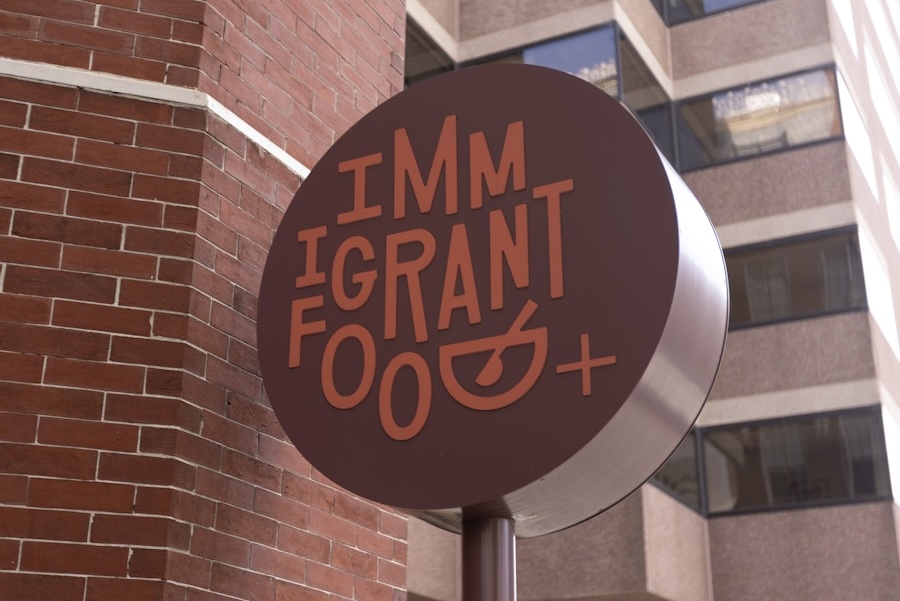 A round outdoor sign for "IMMIGRANT FOOD" with the "D" stylized to look like a bowl and spoon.