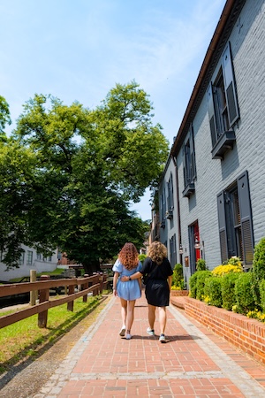 Two women walk down a brick path beside a white building and green trees