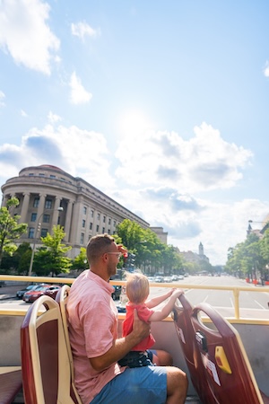 Man holding a toddler on a double-decker bus, city street and blue sky ahead