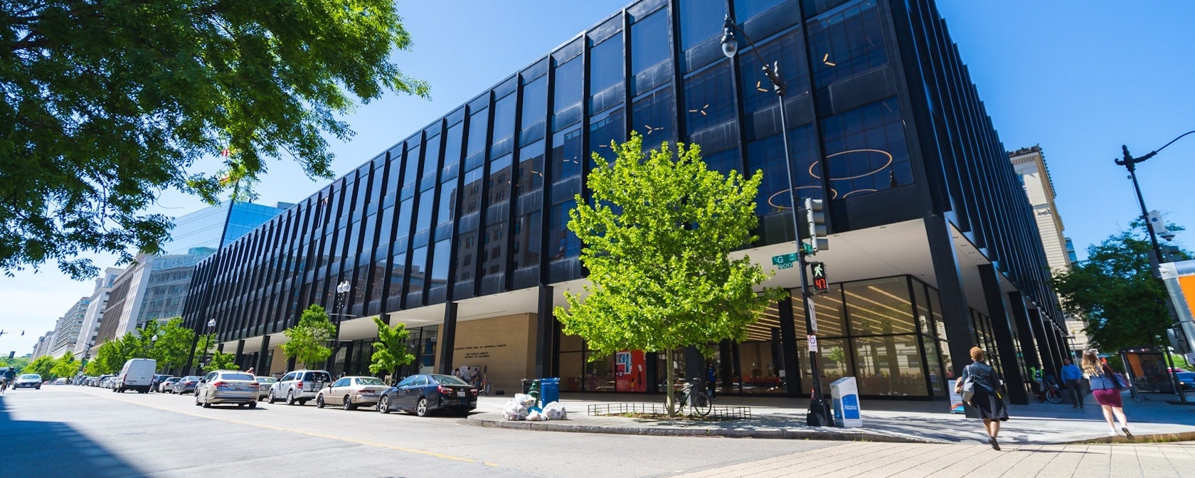 Wide photo of a modern glass office building with trees and street in front