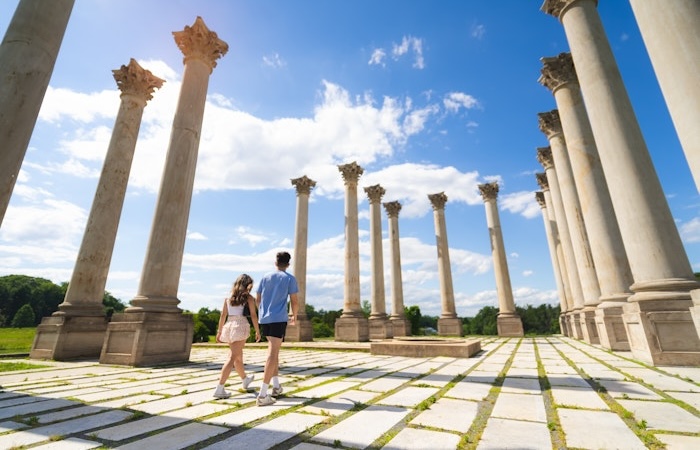Two people walking among tall stone columns under a bright blue sky