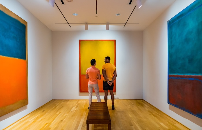 Two people view colorful abstract paintings in a modern gallery with wood floor