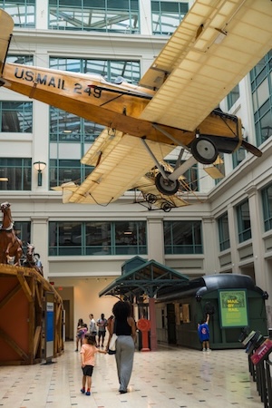 Vintage tan biplane suspended in an indoor atrium above people walking