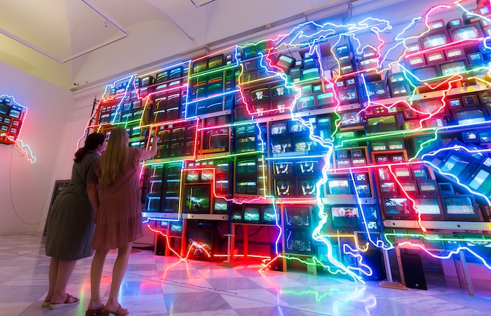 Two people stand in a room facing a large wall of colorful neon circuit-like lights
