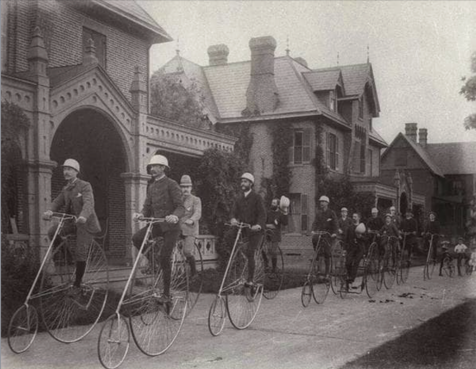 Group of cyclists riding penny-farthings on a village street, black-and-white photo