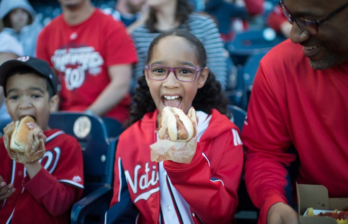 Smiling girl in red Nationals hoodie eats a hot dog at a baseball game