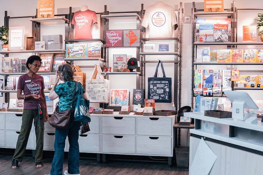 Two women browsing framed art and books in a bright shop interior.