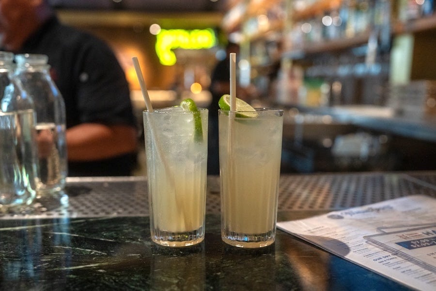Two lime drinks with straws on a bar counter, menu and glass pitcher nearby