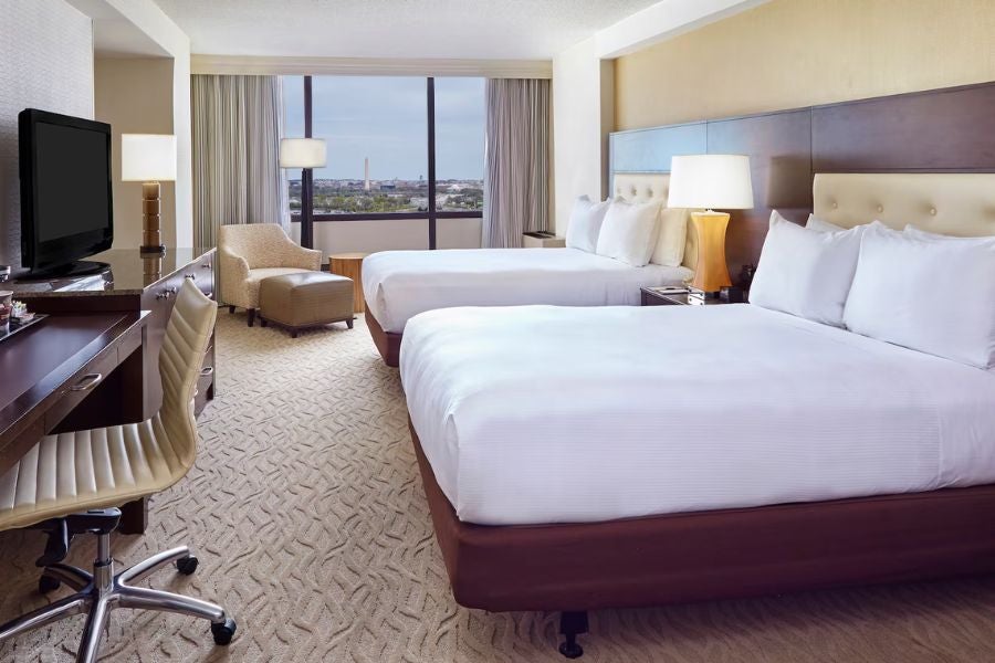 Hotel room with two beds, desk and TV, and large window with city view
