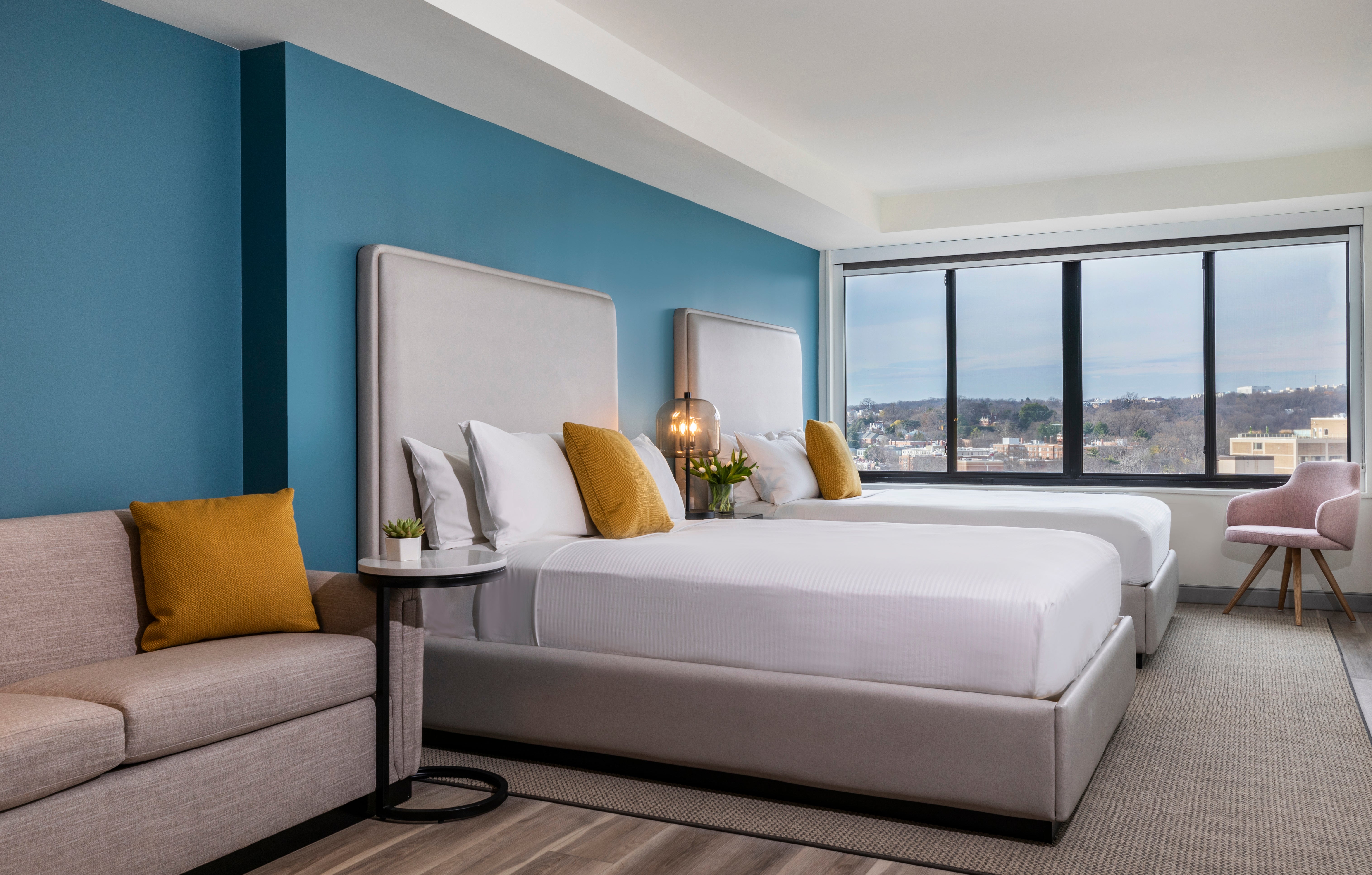 Modern hotel room with king bed, sofa, and floor-to-ceiling windows overlooking city