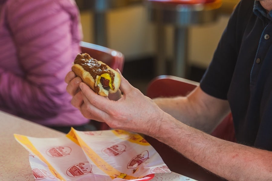 Man holding a chili dog over a tray in a fast-food restaurant.
