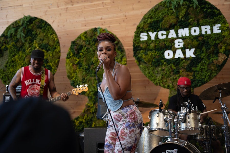 Singer performs on outdoor stage with band; “Sycamore and Oak” sign in greenery behind