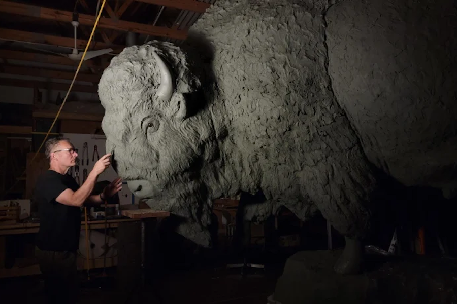 Man in a workshop measuring a large unfinished lion sculpture