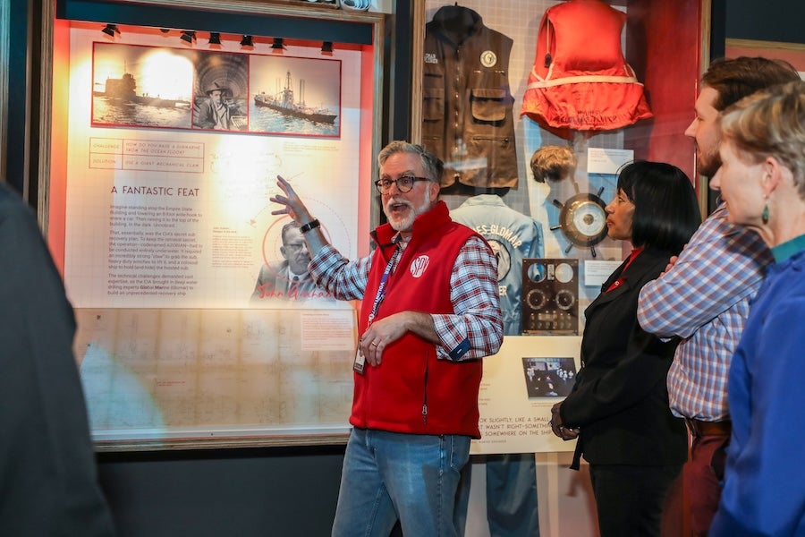 Man in red vest gestures at exhibit text while visitors watch in a museum