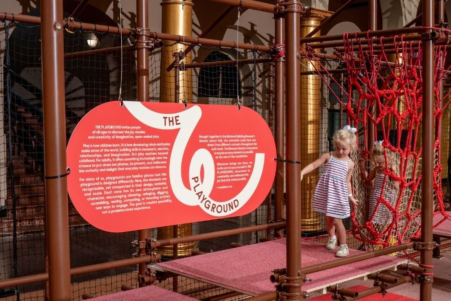 Child exploring a rope climbing structure beside an introductory sign at the National Building Museum’s PLAYGROUND exhibition.