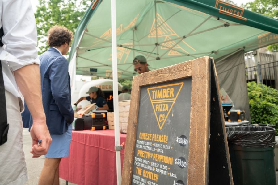 Timber Pizza Co. pop-up stand serving wood-fired pizza beneath a branded canopy, with customers waiting beside a menu board listing pizza offerings.
