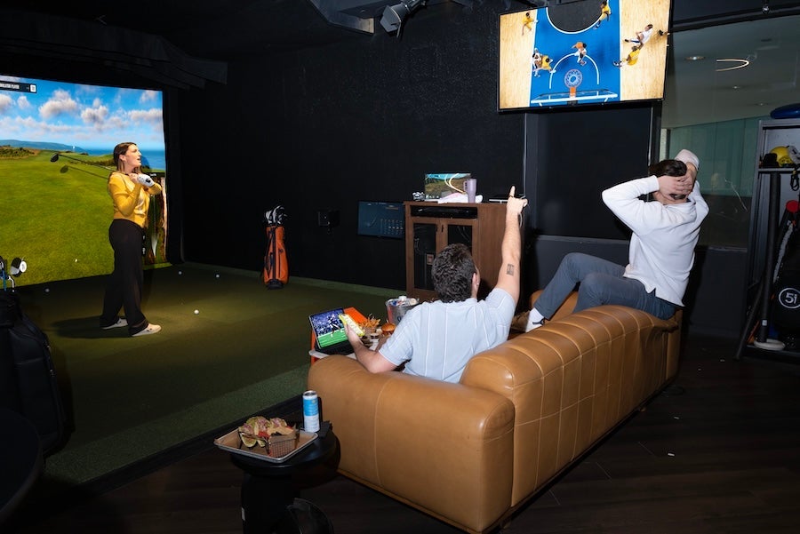 Person swings at indoor golf simulator as another lounges on tan sofa while watching sports.