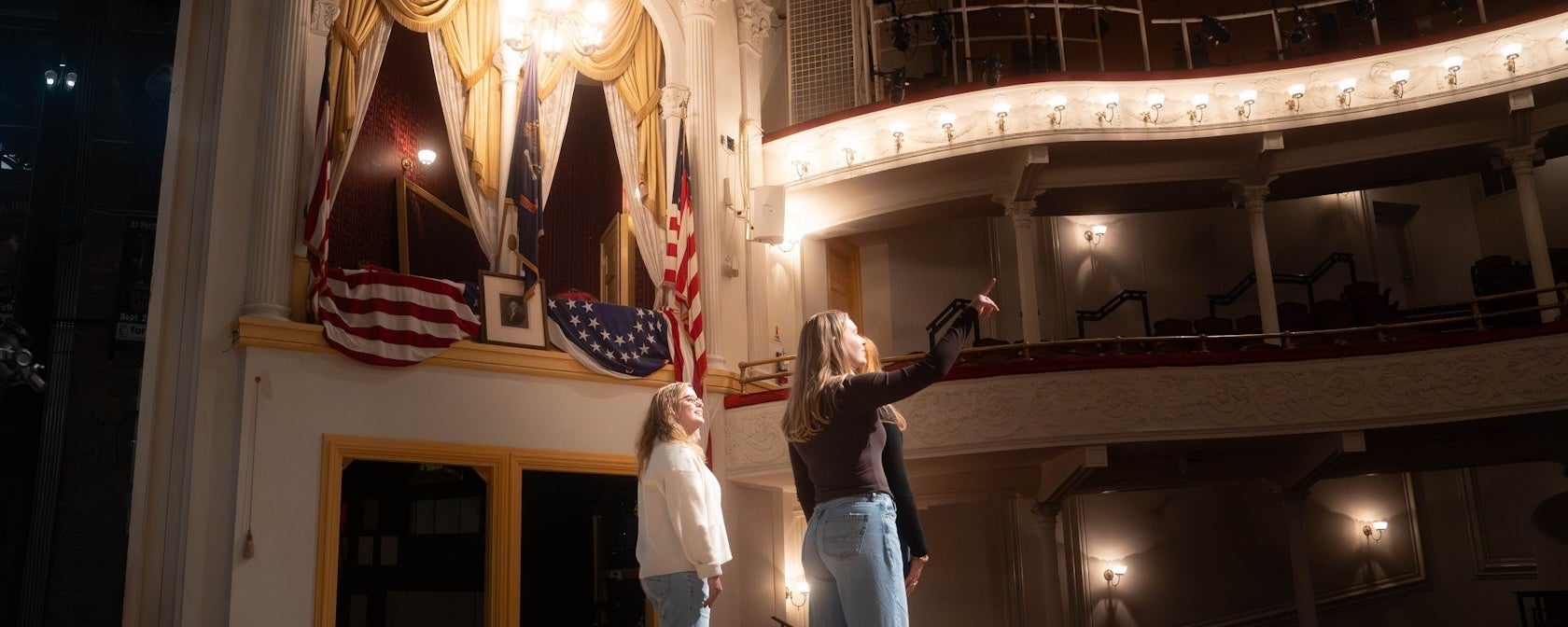 Two performers on stage beneath ornate theater balconies and chandelier.