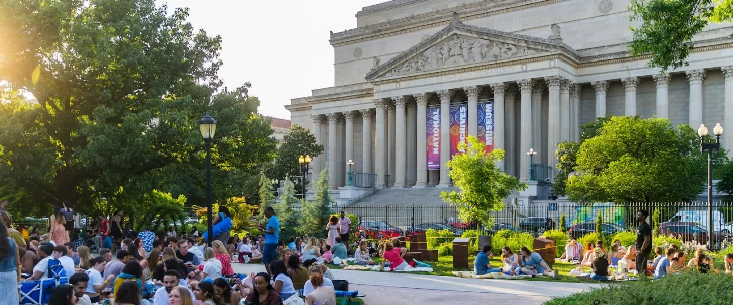 People relax and enjoy a picnic on the lawn during a summer event near the National Archives in Washington, DC, with sunlight filtering through the trees.