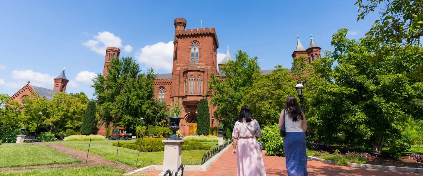 A wide view of the Smithsonian Castle with two people walking toward the entrance through the garden pathway on a sunny day.