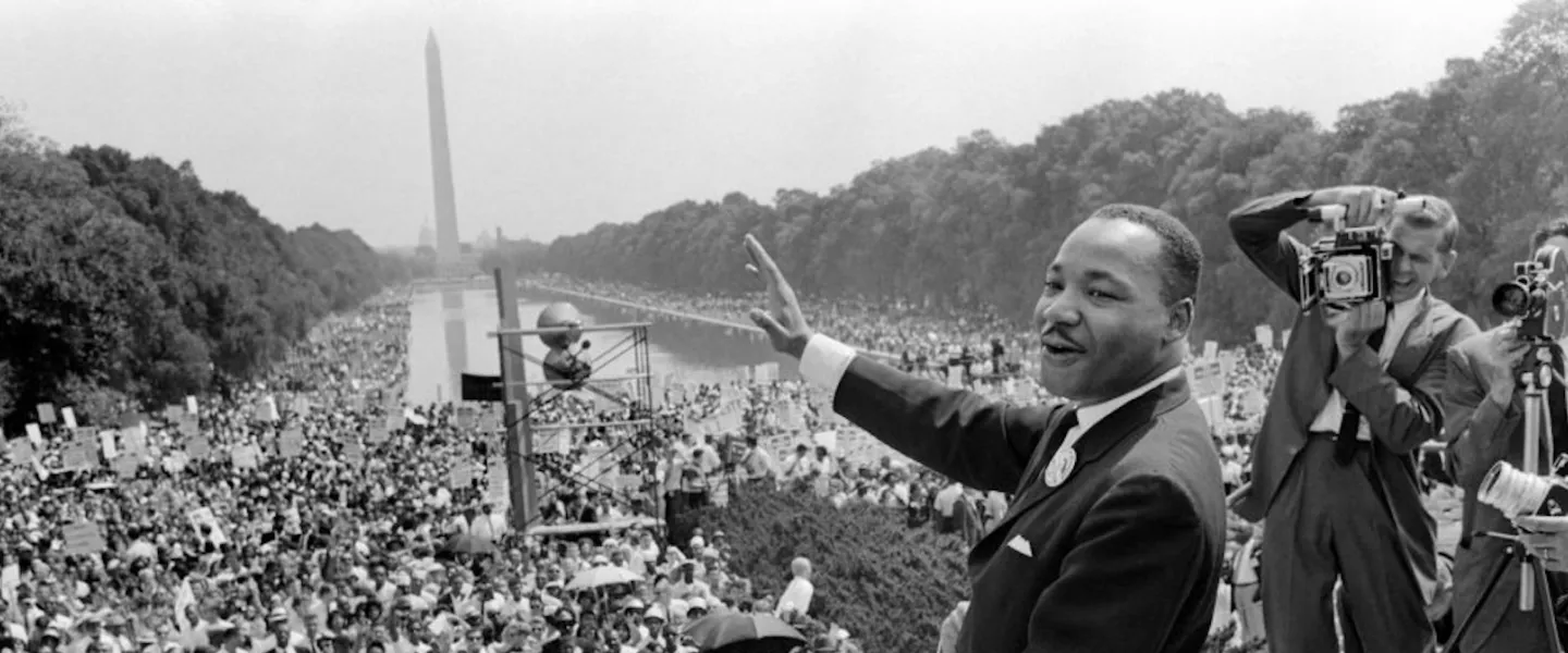 Martin Luther King Jr. waving at crowd during an outdoor speech beside the Reflecting Pool; photographers stand nearby.