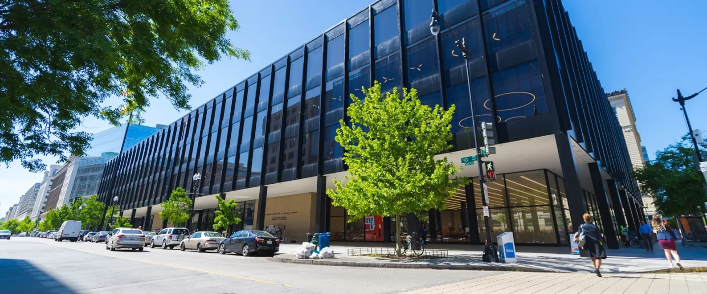 Wide photo of a modern glass office building with trees and street in front